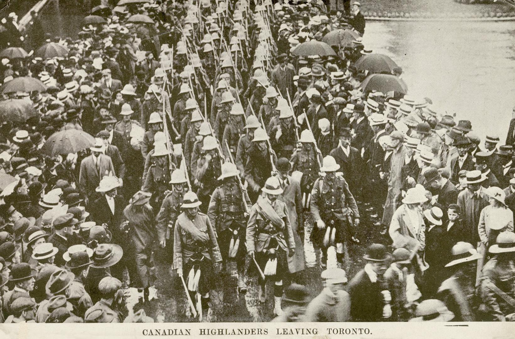 Black and white photograph of soldiers wearing kilts and pith helmets marching on a city road.