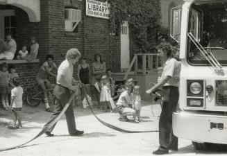 Picture of two fire fighters with hose in front of a crowd of onlookers outside a public librar…