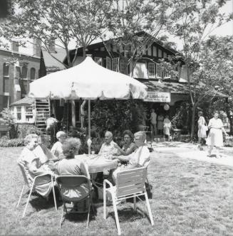 Group of people seated at a table with umbrella at a garden party on library lawn. 