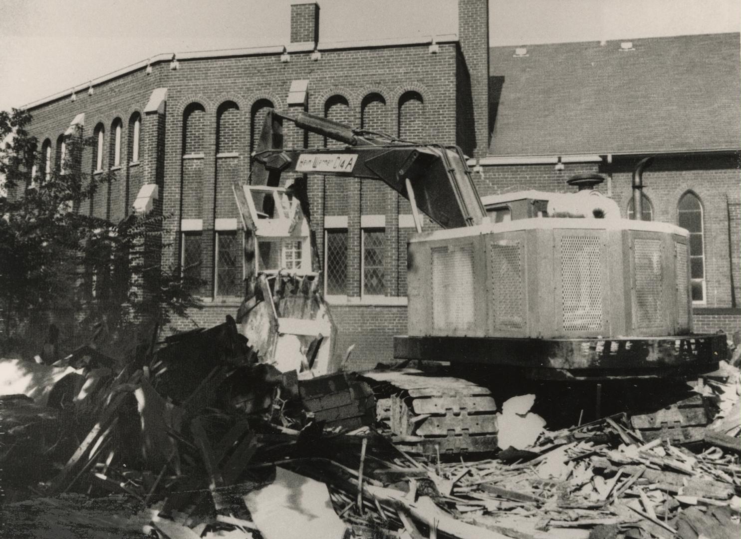 Picture of buldozer demolishing building with other buildings in background. 