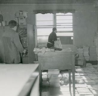 Picture of a room with table and piles of paper and two people working in it. 