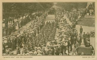 Sepia toned photograph of soldiers marching down a wide street as civilians look on.