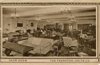 Sepia-toned photo postcard depicting an interior view of a furniture store, showing various sof…