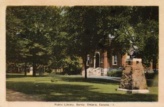 Picture of a brick library building mostly hidden by trees with park and stone memorial in fron…