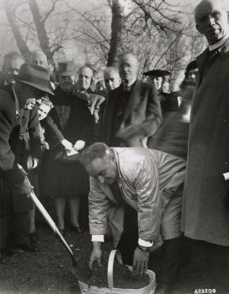 Picture of a man putting soil in a basket while others stand and watch at sod turning ceremony.…