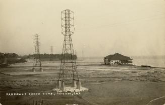 Black and white photograph of a large building built on a shoreline. Electrical transmission to…