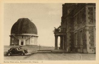 Black and white photograph of two cars parked outside a stone building. Domed building in the b…