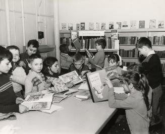 Picture of a group of boys and girls reading books at a large table while others look at books …