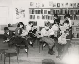 Picture of a group of boys and girls reading books while sitting on stools while others look at…