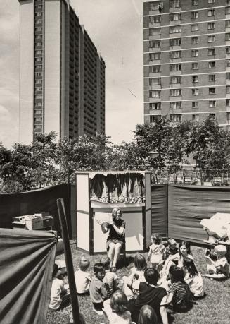 Picture of children sitting outside listening to a librarian storyteller in a park with apartme…