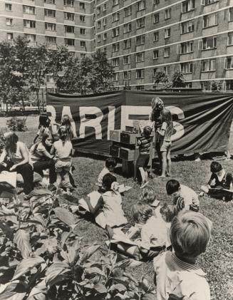 Picture of a group of children on a lawn looking at books with apartment building in background…