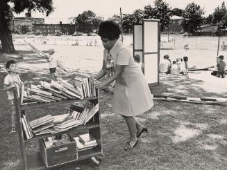 Picture of a group of children on a lawn looking at books with apartment building in background…