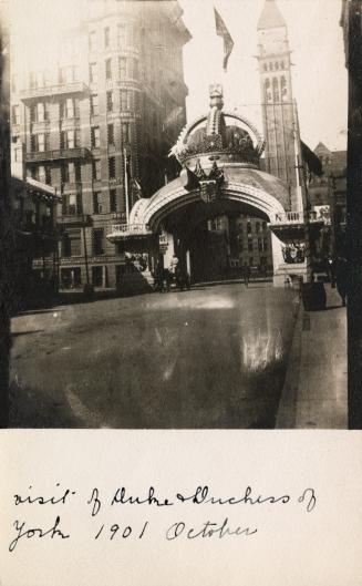 Black and white photograph of an arch with a gigantic crown on the top of it spanning a city st…