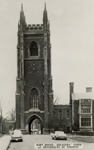 Black and white photograph of a bell and clock tower with a gothic archway providing access thr…