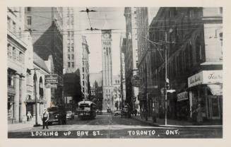 Black and white photograph of a city street with skyscrapers on either side and a clock tower a…