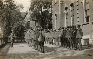 Black and white photograph of soldiers lined up for role call in front of a collegiate building…