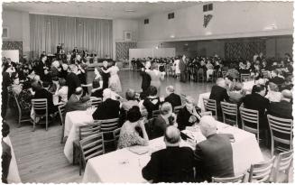 Black and white photo postcard depicting a ballroom with a band on stage and several couples da…