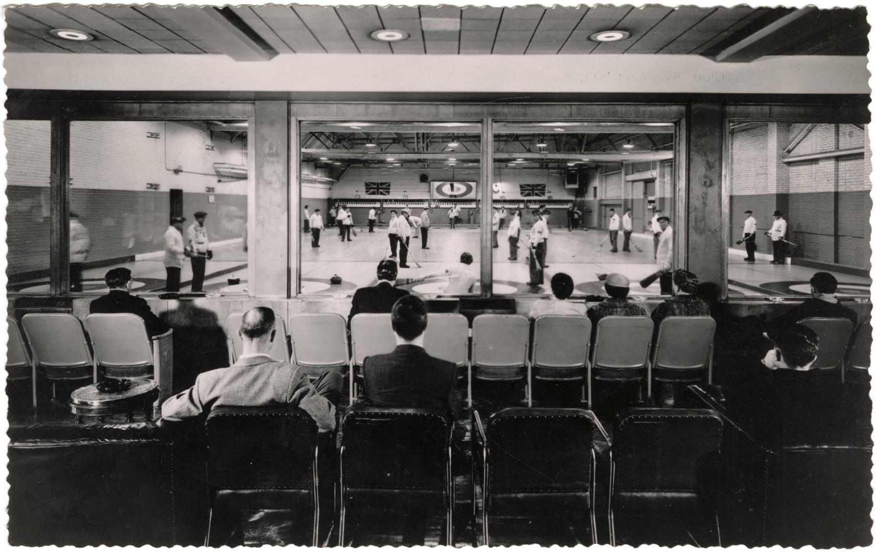 Black and white photo postcard depicting a room of men engaged in curling while onlookers watch…