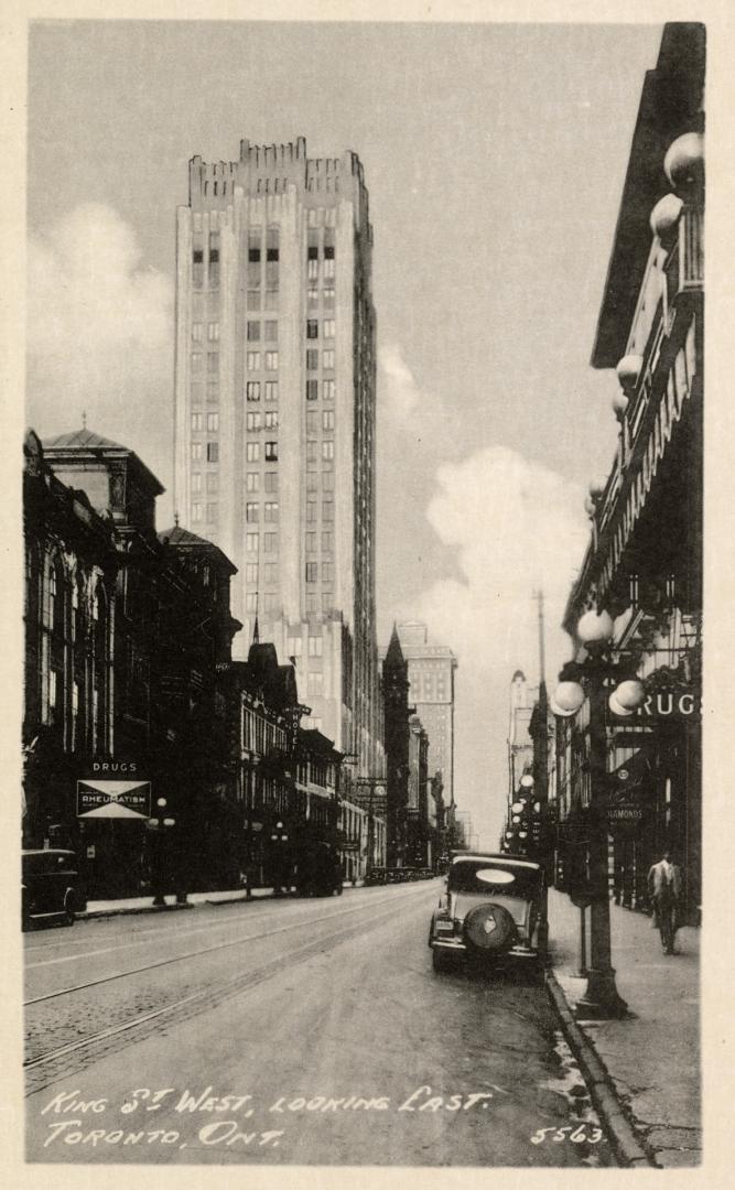 Black and white photo postcard of King Street looking east with a car and shops in view. A shop…