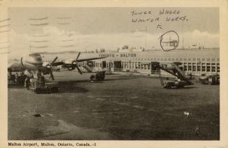 Black and white photo postcard depicting an airplane in front of a flat, wide building with an …