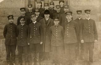 Black and white photo postcard depicting a group of 16 men in uniform with caps. A man in the c…