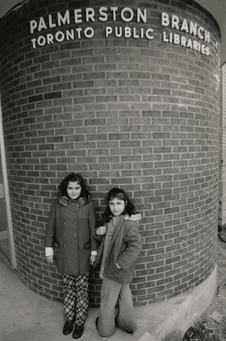 Two girls stand outside Palmerston Branch library with library sign above them. 