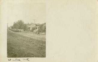 Black and white postcard depicting an inset photo of a dirt road with many men trailing behind …