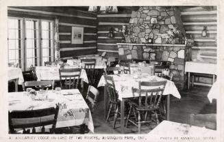 Black and white photograph of tables set for four people in a room in log cabin with fieldstone…