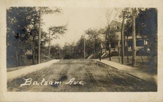 Black and white photo postcard depicting a view down an unpaved residential street with a large…