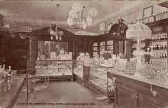 Brown-toned photo postcard depicting the interior of a shop with a man standing in front of a c…