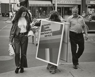 Person wearing a sandwich board advertising library hours stands on the street with two people …