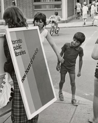 Person wearing a sandwich board advertising library hours stands on the street with boy looking…