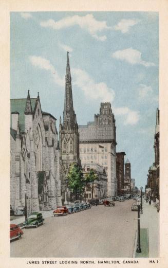 Colorized photograph city street with parked cars lining it. Churches are on the left hand side…