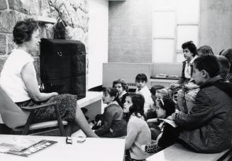 Picture of interior of library with a staff member sitting in a chair speaking to a group of ch…