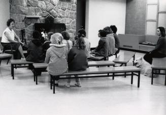 Picture of interior of library with a staff member speaking to a group of children sitting on b…