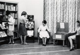 Picture of interior of library. Two women standing looking a vinyl records and two women wearin…