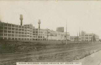 Black and white photo postcard depicting the 3-4 large warehouse-style buildings. The caption a…