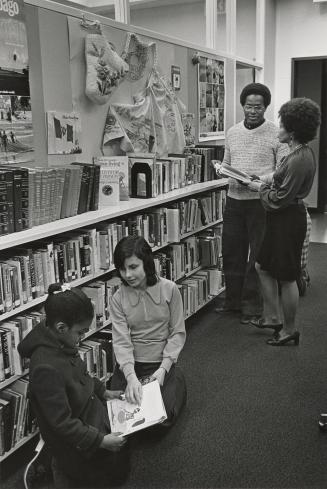 Picture of two library staff standing near shelves and two children sitting on the floor lookin…