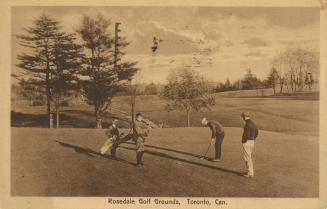 Sepia-toned postcard depicting a golf course with four men in the forefront playing golf. The c…