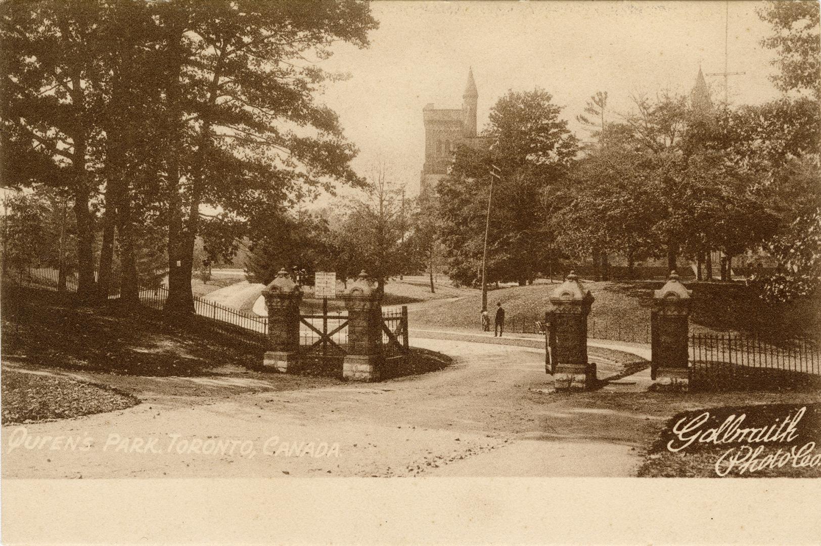 Black and white photograph of an open gate in an iron rail fence. In the background are large,  ...